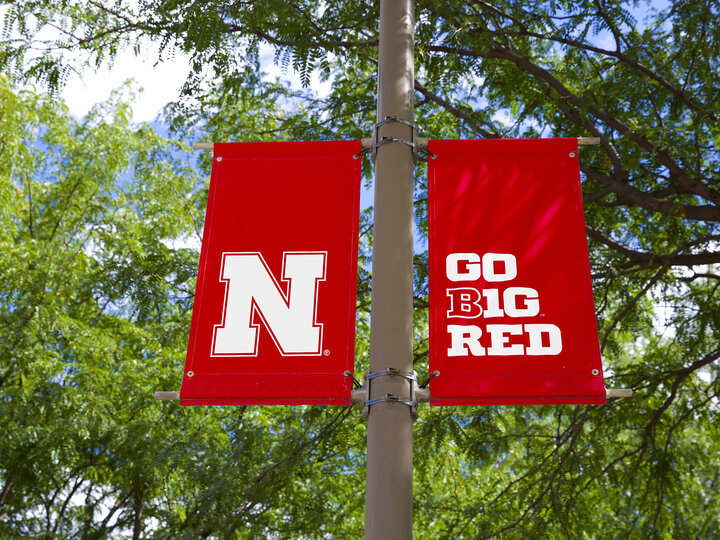 White Nebraska N logo and Go Big Red graphic on red light pole banners with trees in the background