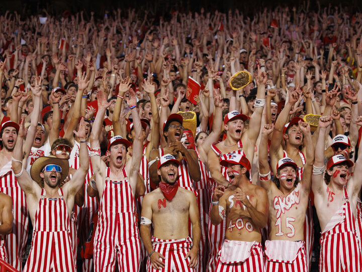 A group of fans in red and white striped overalls cheer the Huskers at a nighttime football game