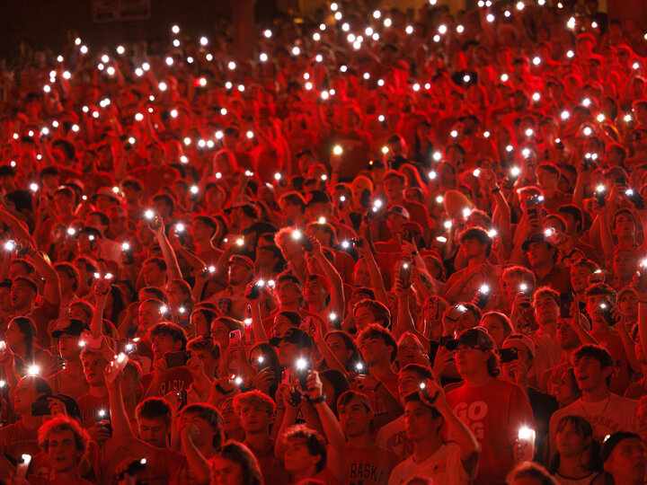 At a nighttime Nebraska football game, the crowd, some holding up phone flashlights, is bathed in red light from above