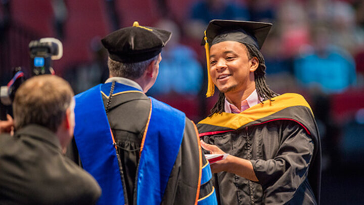 A graduate in full regalia accepts his diploma and shaking the chancellor's hand.